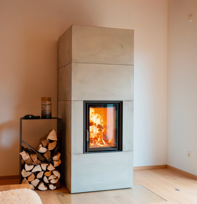 „Moderner Kaminofen mit hoher, schlanker Bauform in einem Wohnraum, zeigt effizientes und emissionsarmes Heizdesign; im Hintergrund gut organisierter Holzstapel und integriertes Regal für Smart-Home-Komponenten zur Steuerung der Heizanlage. - © Foto: Brunner